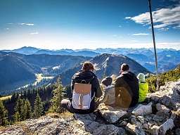 Familie sitzt auf einem Berggipfel mit Blick auf Tannenwälder und Gipfel